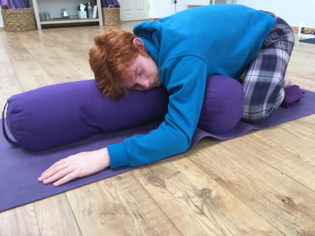 Man lying on a yoga mat in supported child's pose using two bolsters as props
